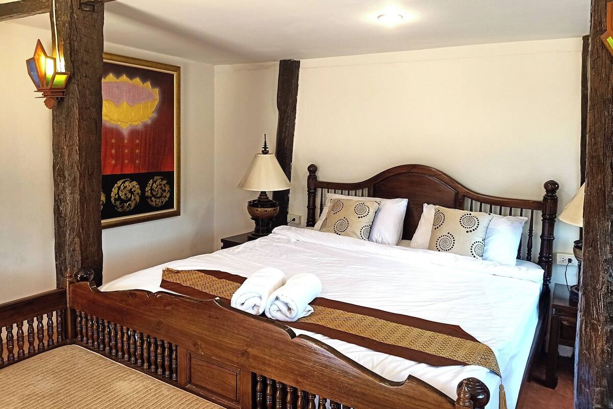 Una cama con estructura de madera, ropa de cama blanca, una lámpara y almohadones decorativos.