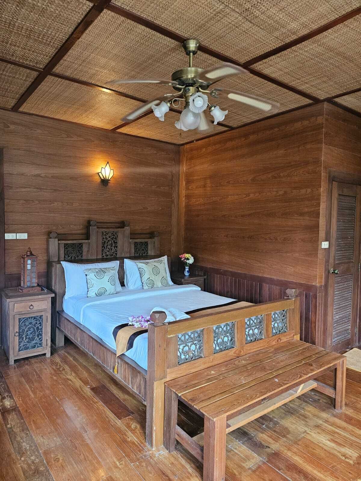 Een slaapkamer met houten details, een bed, nachtkastjes, een bankje, een plafondventilator en een wandlamp.