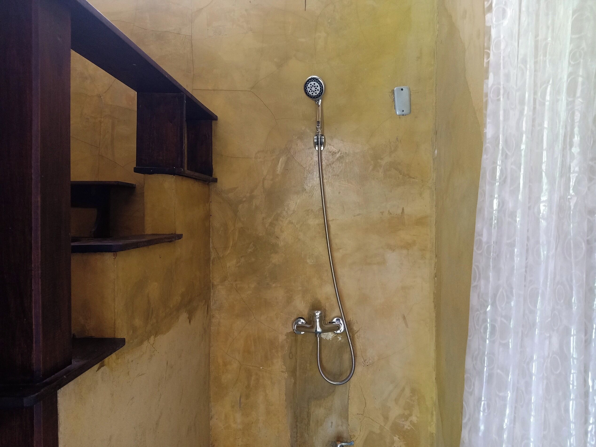 Une douche équipée d’un pommeau de douche à main et d’un robinet mural, dans une pièce dont un mur est jaune et présente une texture particulière, et qui est également dotée d’étagères en bois.