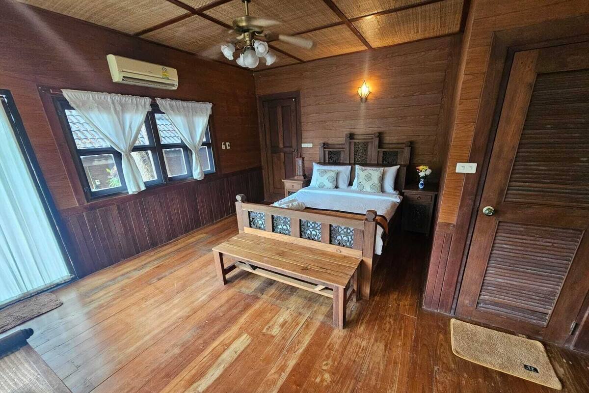 Una habitación de madera con una cama, un banco, un ventilador de techo y una ventana con cortinas.
