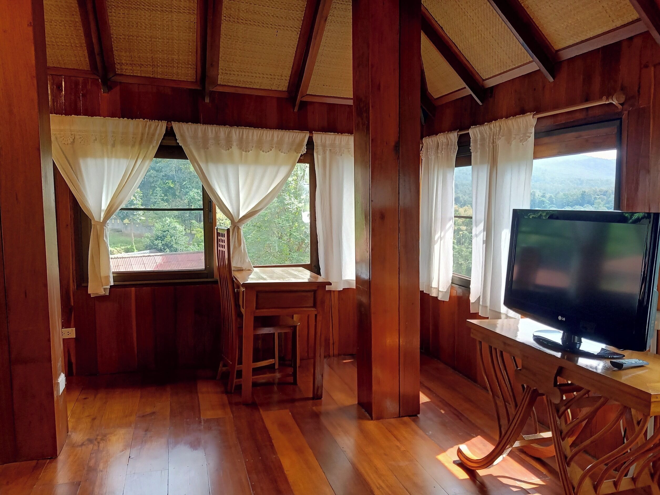 Un interior de madera con un televisor sobre una mesa de madera, una silla de madera y una ventana con cortinas.