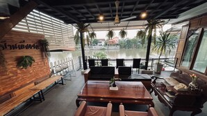Un restaurante con interior de madera, una mesa grande y sillas. Hay plantas y se ve una masa de agua desde el exterior.