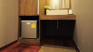 Un pasillo estrecho con piso de madera, un refrigerador pequeño, un lavamanos con un espejo encima y una ventana con cortinas.