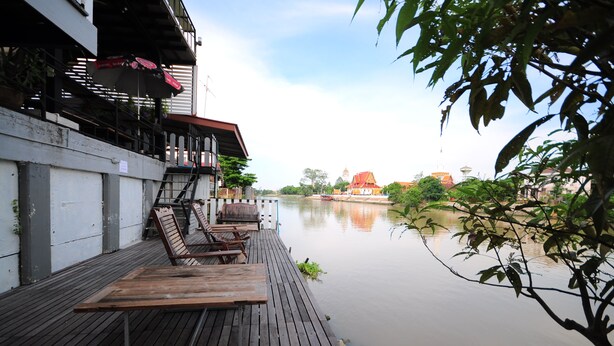 Una terraza de madera con sillones de descanso con vista a un río tranquilo, un edificio con techo rojo al fondo y una rama de árbol en primer plano.