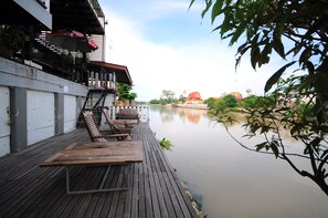 Una terraza de madera con sillones de descanso con vista a un río tranquilo, un edificio con techo rojo al fondo y una rama de árbol en primer plano.