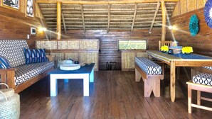 Um interior rústico de madeira com teto alto, luminárias de palha no teto e uma área de estar central com almofadas estampadas e uma mesa.