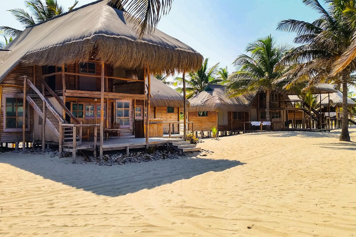 A beachside wooden building with a thatched roof, a staircase leading to the entrance, and a sandy beach in the background.