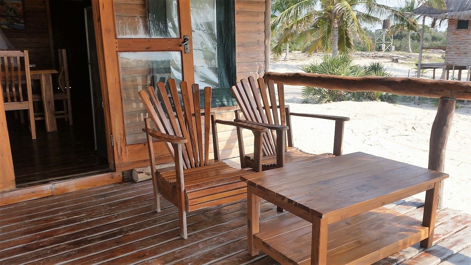 Una terraza de madera con una mesa y dos sillas, una puerta de vidrio que da al interior, y vistas de palmeras y una playa al fondo.