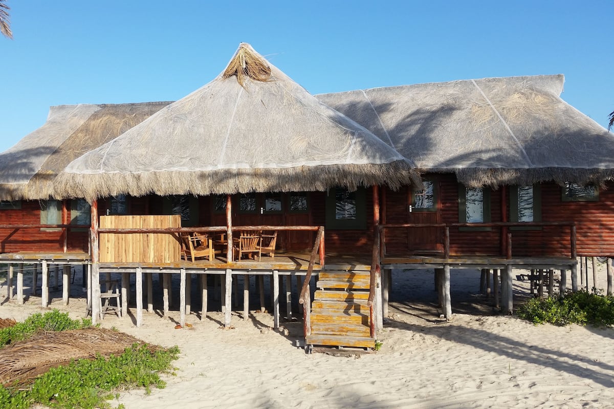 A wooden structure with thatched roofs, a wooden deck, and a small table with chairs.