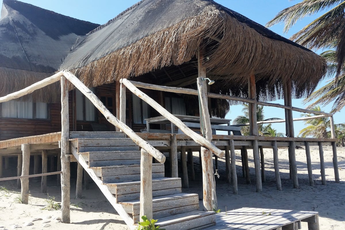 A wooden structure with a thatched roof, wooden stairs, and a sandy beach in the background.