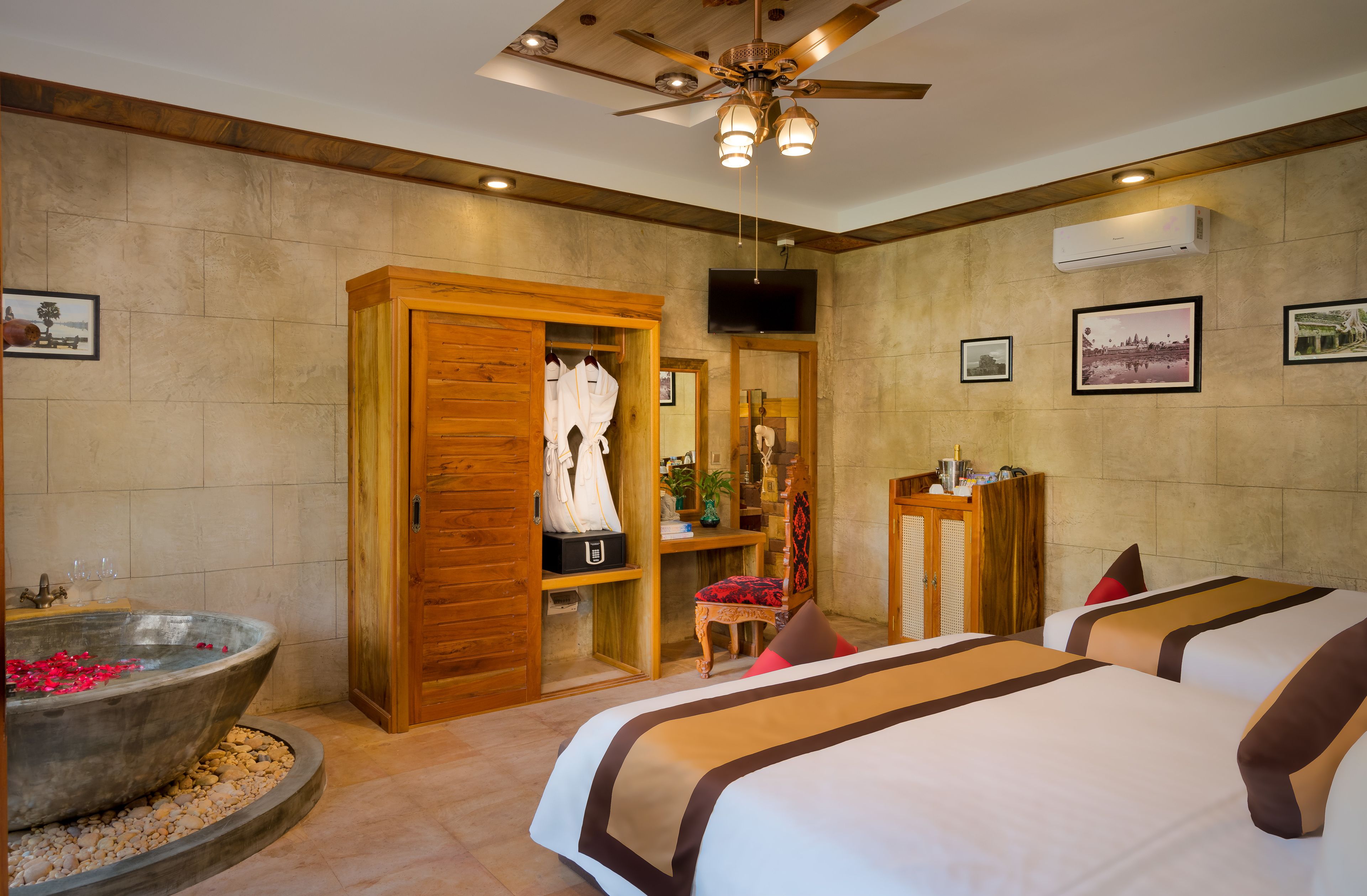 A hotel room with a large wooden wardrobe, a bathtub with rose petals, two beds with brown and white bedding, a ceiling fan, and framed pictures on the wall.