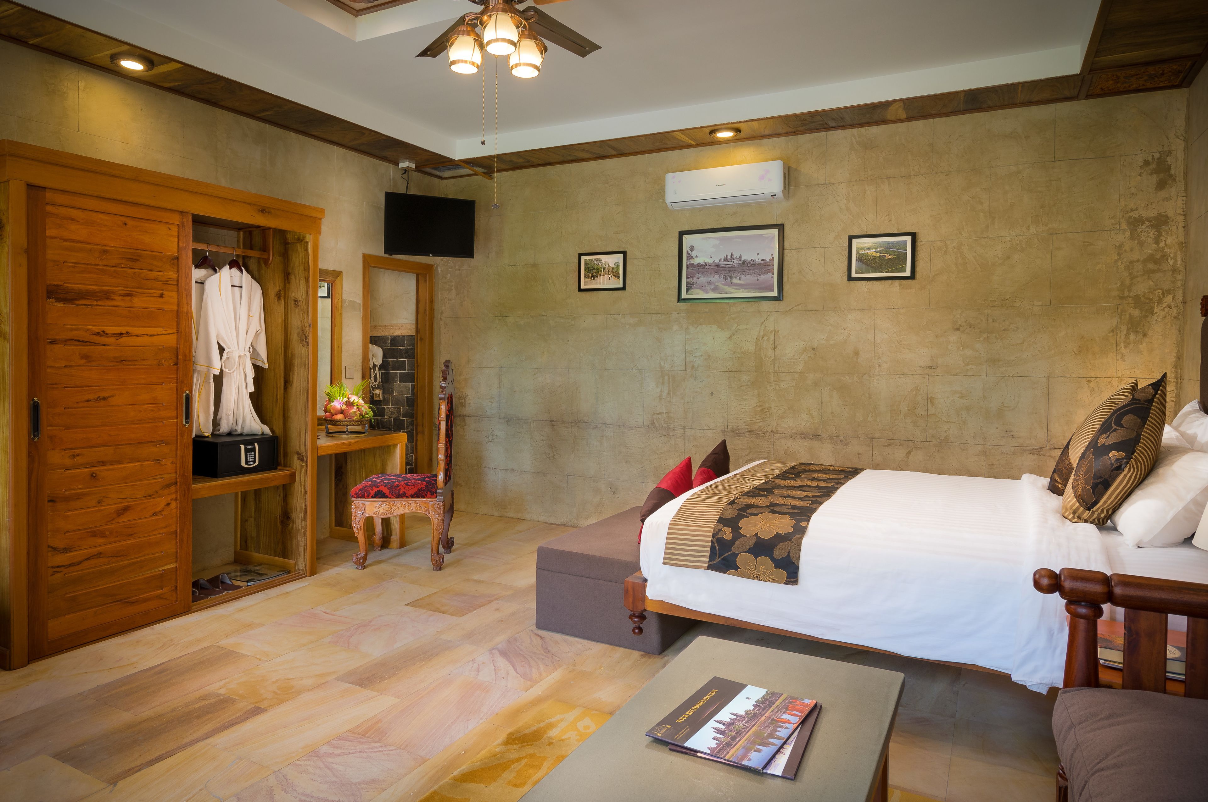 A hotel room with a bed, a wooden wardrobe, a small table with a magazine, a chair, and a ceiling fan.