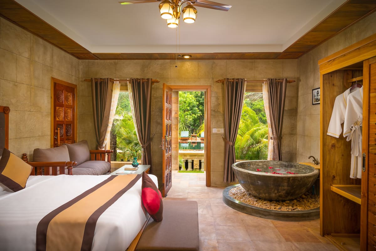 A spacious bedroom with a large bed, a sitting area, a bathtub, and a view of a lush garden.