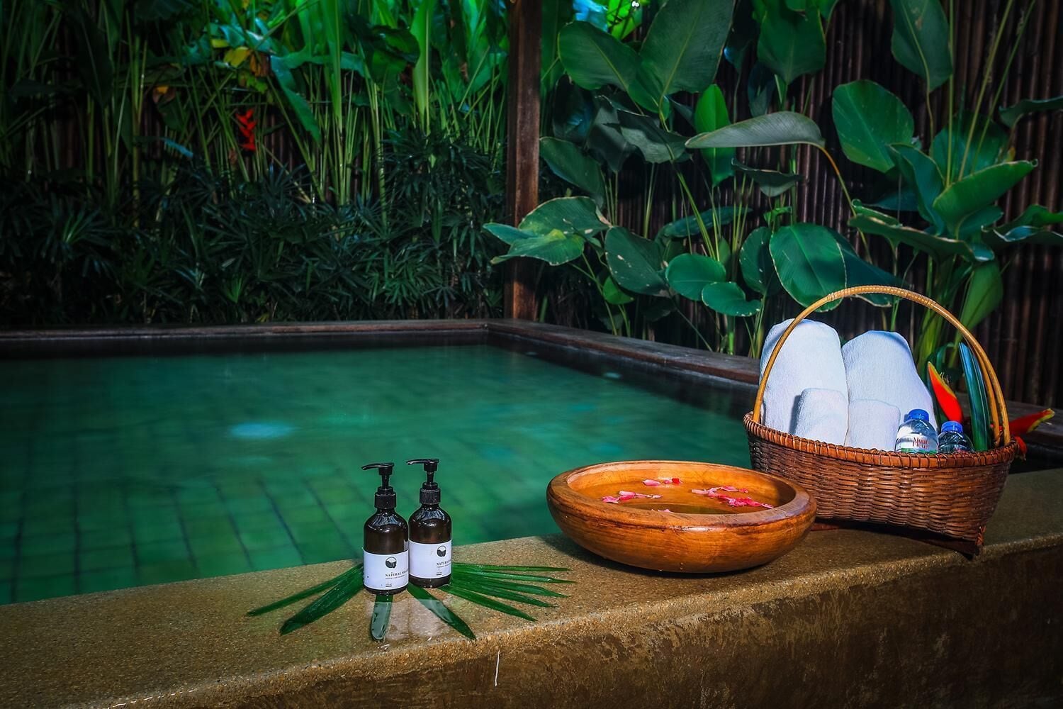 A poolside setting with a wooden bowl, two bottles, and a basket containing towels and toiletries.