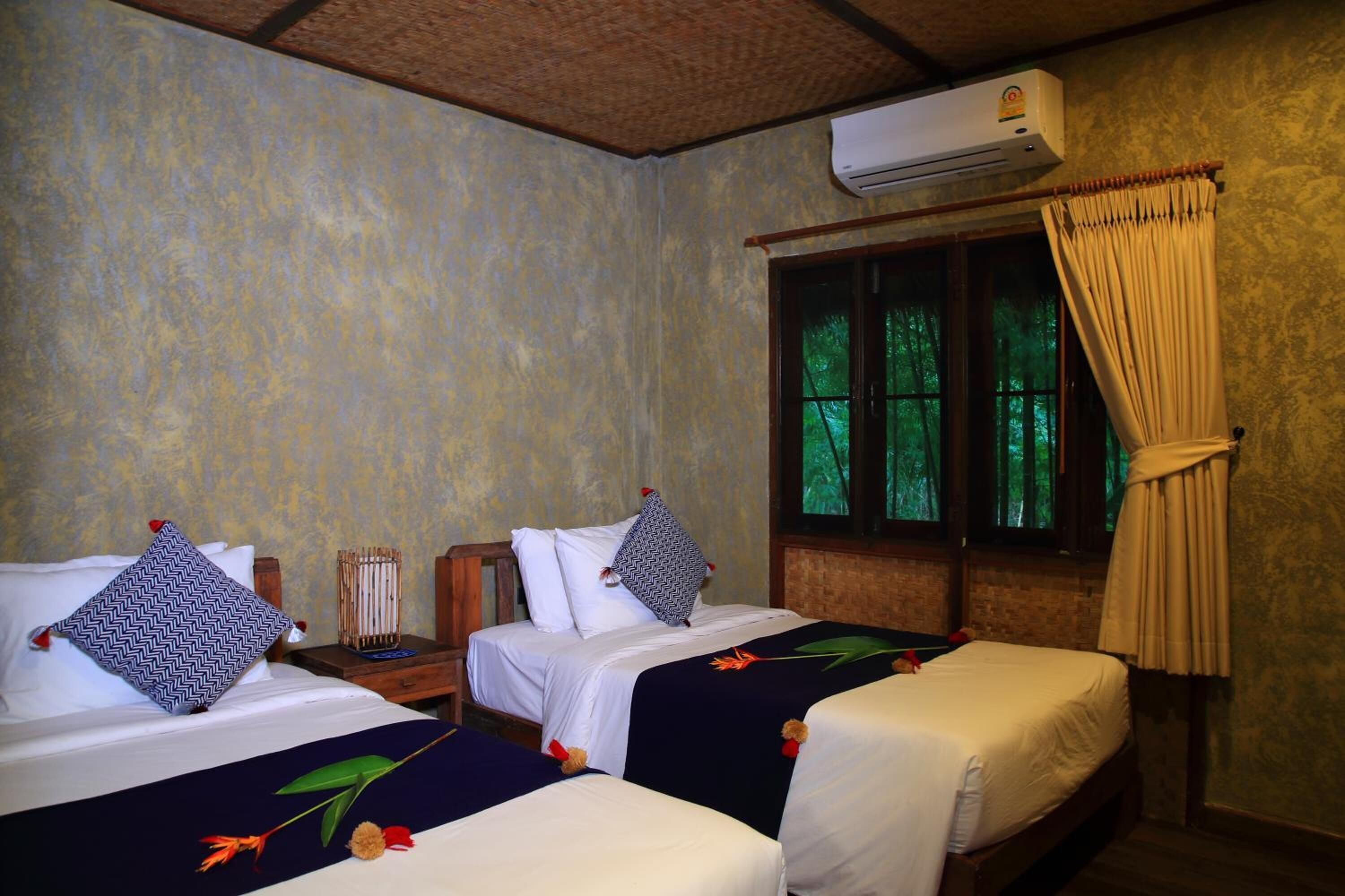 A room with two beds, each with a patterned pillow and a floral bedspread. There is a window with curtains, an air conditioning unit above the window, and a wooden headboard.