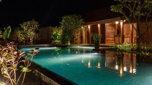 A nighttime view of a swimming pool with wooden deck and lounge chairs, surrounded by lush greenery and wooden structures. - Azalea Village Wellness Resort (Chiang Dao)