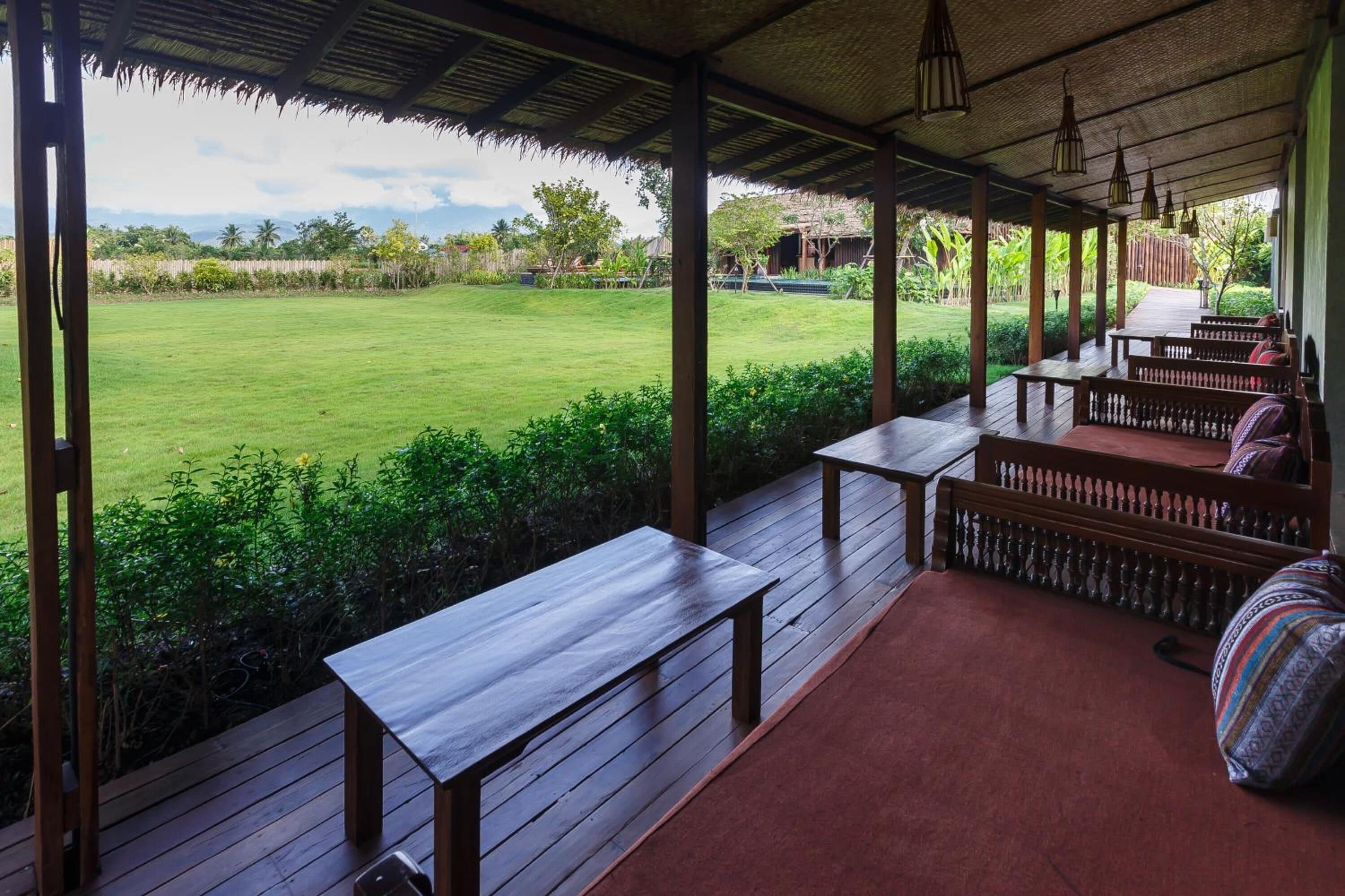 An outdoor covered area with wooden benches and tables, a thatched roof, and a view of a grassy field.