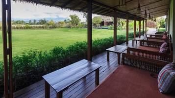 An outdoor covered area with wooden benches and tables, a thatched roof, and a view of a grassy field.