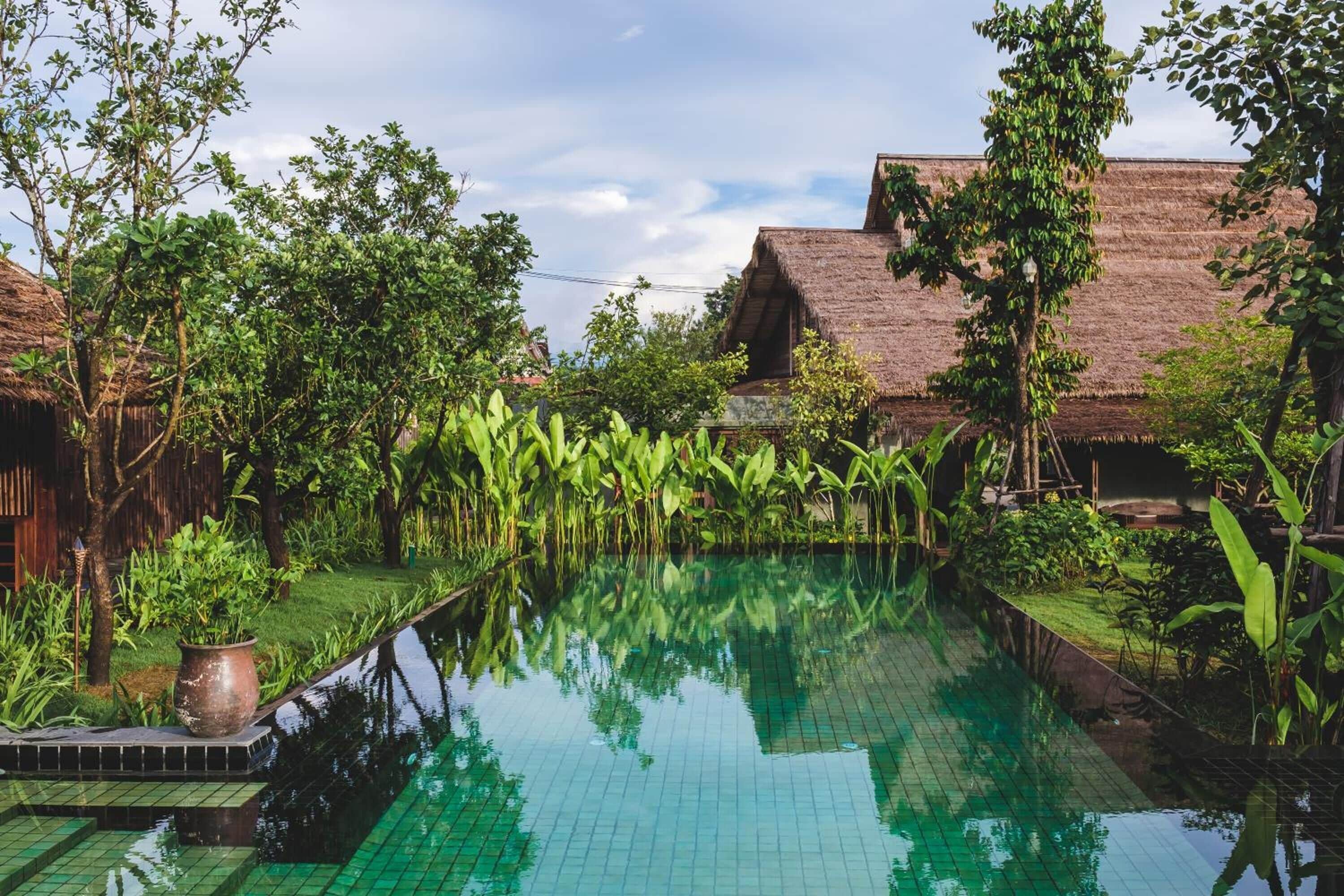 Una piscina rodeada de exuberante vegetación y edificios tradicionales con techo de paja.