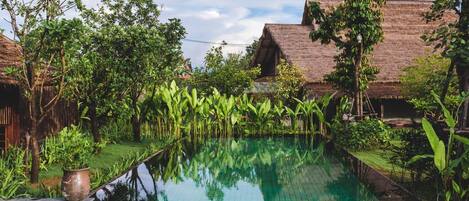 Una piscina rodeada de exuberante vegetación y edificios tradicionales con techo de paja.