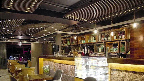 Un restaurante con mesas y sillas de madera, una zona de bar con estanterías llenas de botellas, y una vitrina con alimentos.