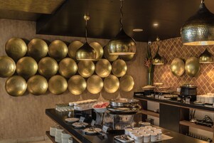 A buffet area with a wall of golden bowls, a variety of food items, and a counter with plates and cups.