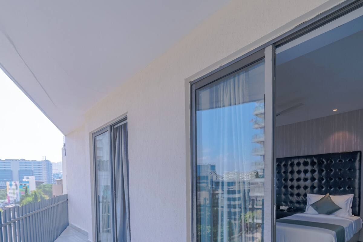 A modern hotel room with a balcony, a sliding glass door leading to a city view, and a neatly made bed with pillows.