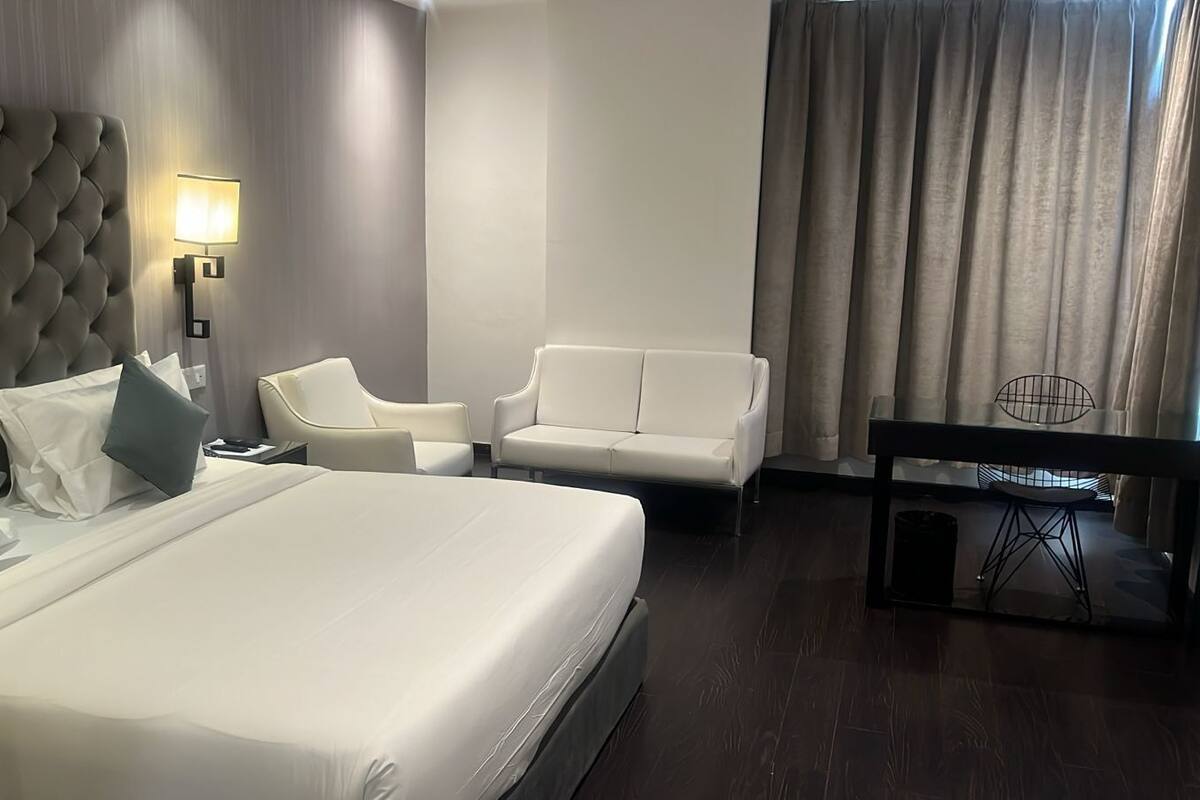 A modern hotel room with a large bed, two white sofas, a small table, and a wall-mounted lamp.
