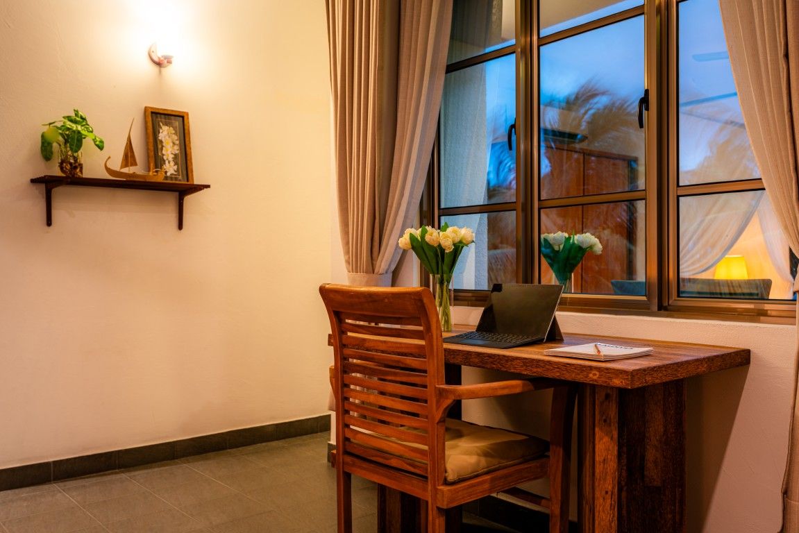 Un escritorio de madera con una computadora portátil y un jarrón de flores, una silla, una estantería con una planta y un cuadro, y una ventana con cortinas.