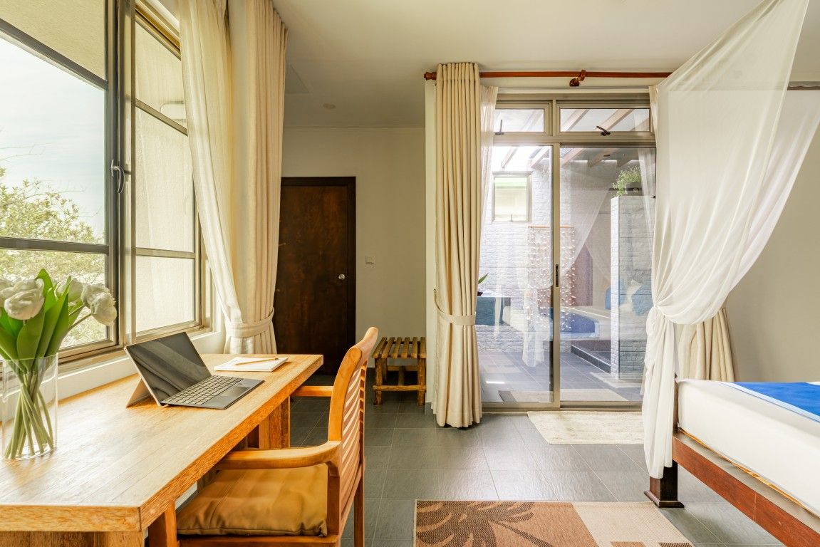 A room with a wooden desk, a laptop, a chair, a bed, and a view through a glass door.