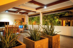 Ampla sala de estar com teto alto, vigas de madeira, uma grande janela com vista para plantas tropicais e um espaço de estar com mesa e cadeiras.