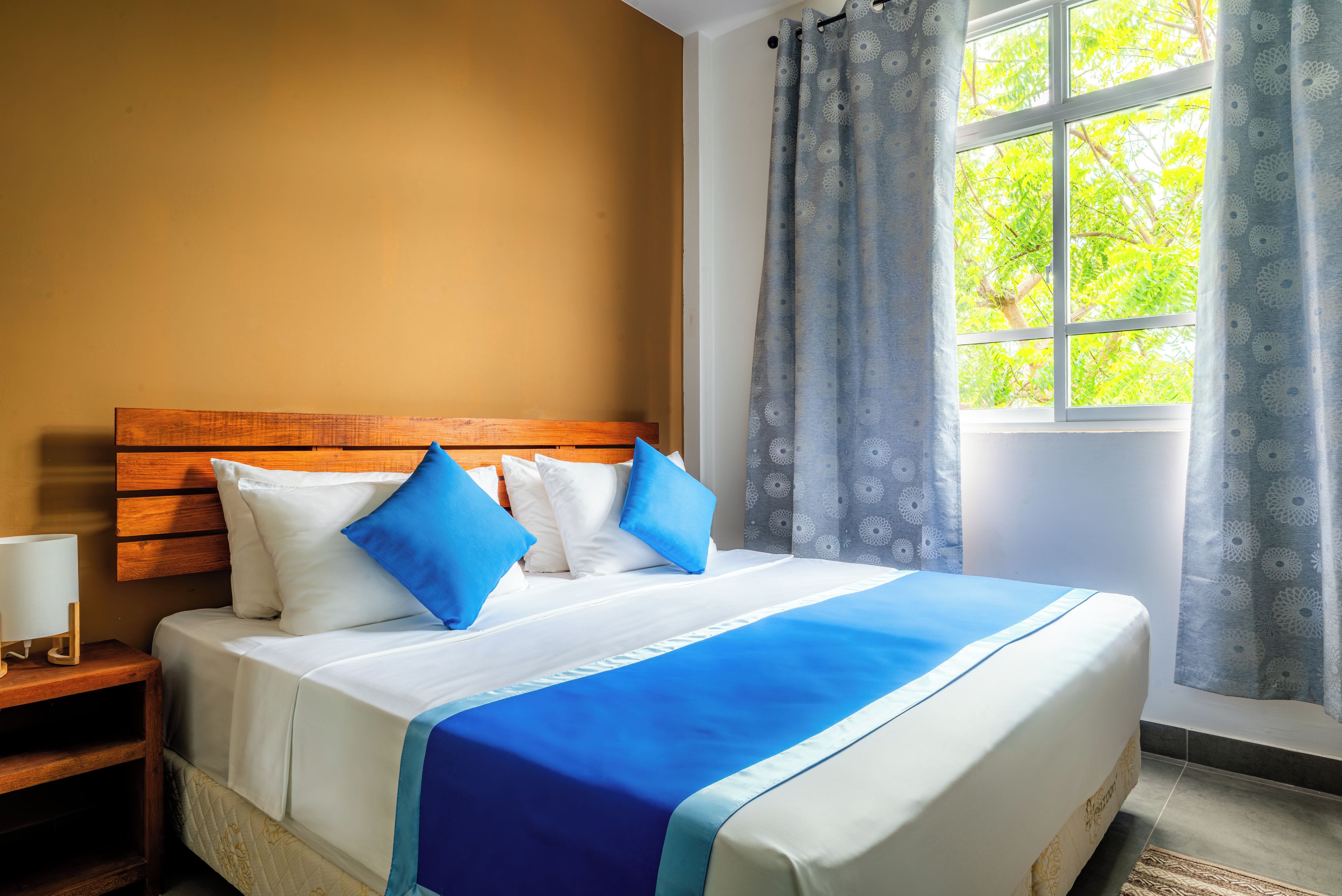Habitación de hotel con cabecera de madera, cama con ropa de cama blanca y azul, mesita de noche con lámpara y ventana con cortinas.