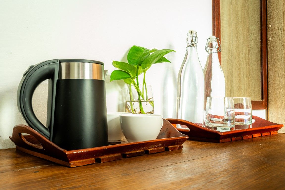 Una tetera de acero inoxidable sobre una bandeja, un cuenco blanco, una planta en maceta y dos botellas de vidrio sobre una mesa de madera.