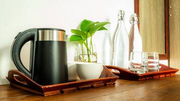 Una tetera de acero inoxidable sobre una bandeja, un cuenco blanco, una planta en maceta y dos botellas de vidrio sobre una mesa de madera.