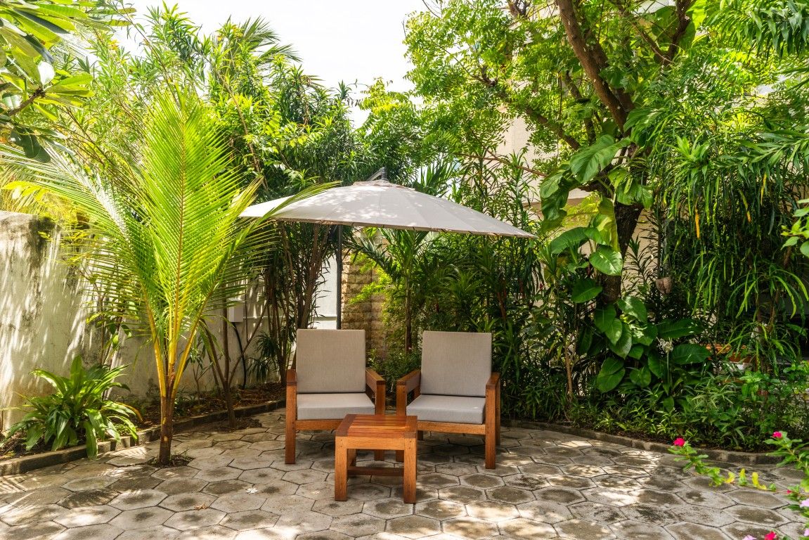 Un área de estar al aire libre con dos sillas y una mesa bajo una pérgola, rodeada de abundante vegetación.