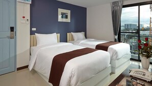 A hotel room with two beds, a large window with a view of the city, a blue door, and a bedside table with a phone and magazine. - Casa Residence Hotel (Bangkok)
