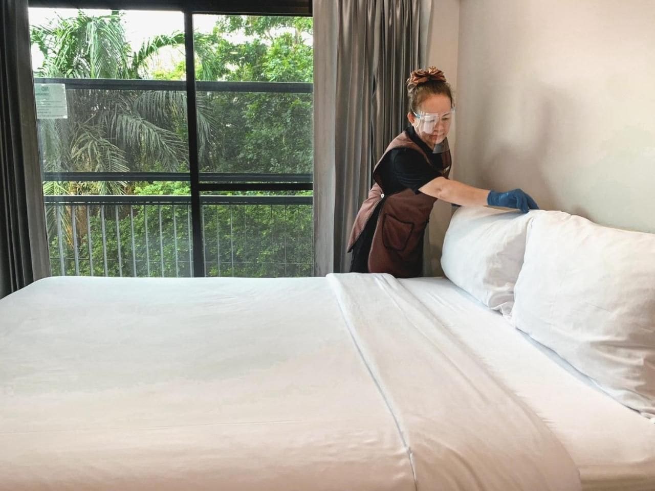 Ein Hotelzimmer mit einem großen Bett, einem Fenster mit Blick auf Grünflächen und einer Person, die die Bettdecke zurechtzieht.