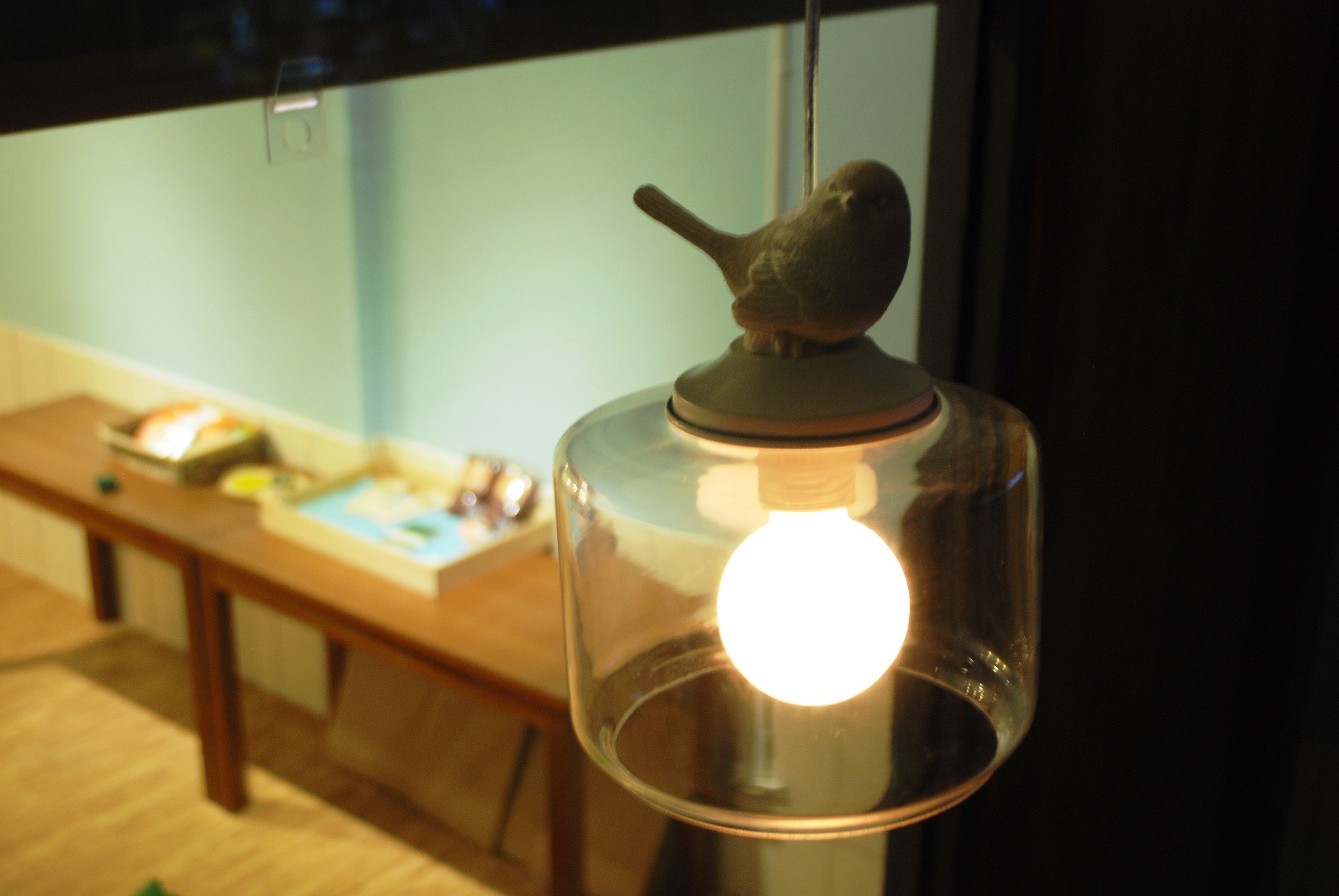 A bird figurine on a lamp with a clear glass enclosure, a wooden table with books and a lamp in the background.