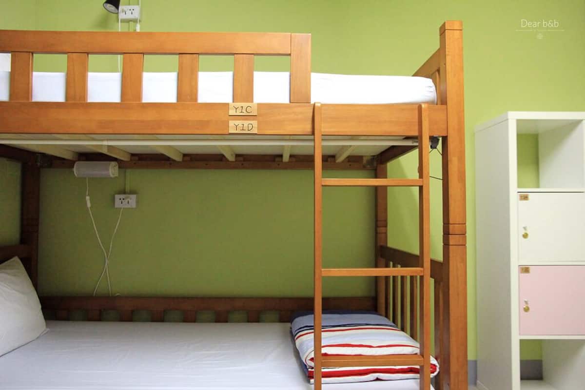 Una cama litera con una escalera, una mesita de noche con lámpara y un armario con varios compartimentos.