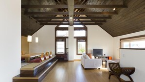 A spacious loft with a high ceiling, wooden beams, a dining area with a long table and benches, a sofa, a TV, and a large window providing natural light. - Ginsetsu (Kutchan)