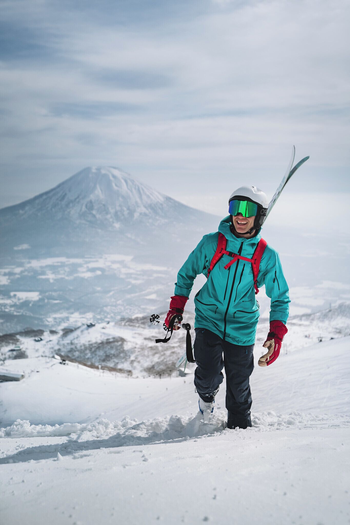 A person in a teal jacket and red gloves walking on a snowy path with a mountain in the background.