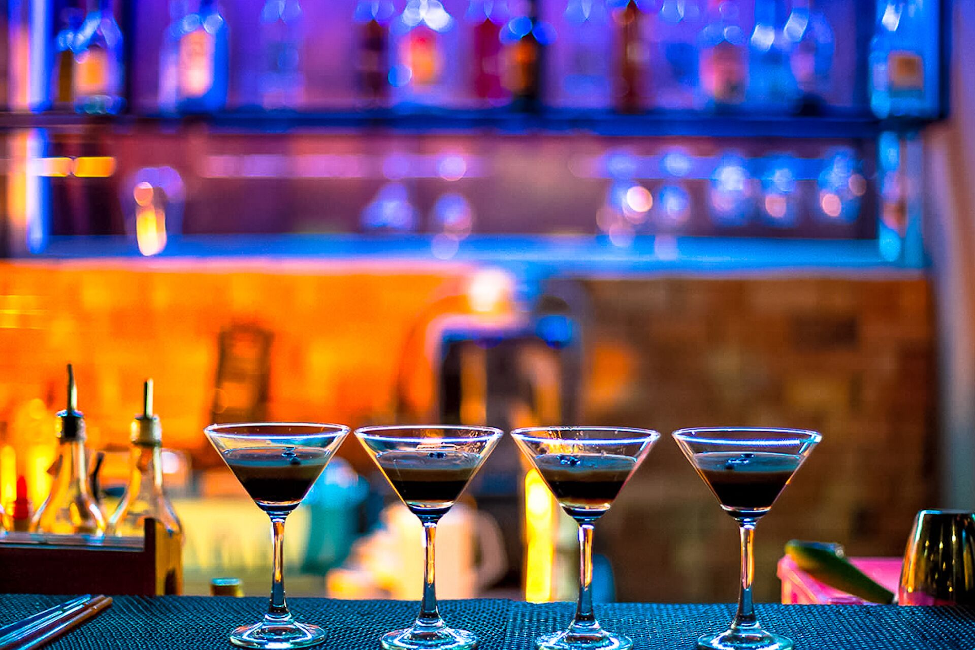 A bar setting with four martini glasses on a table, bottles in the background, and a blurred bar area.