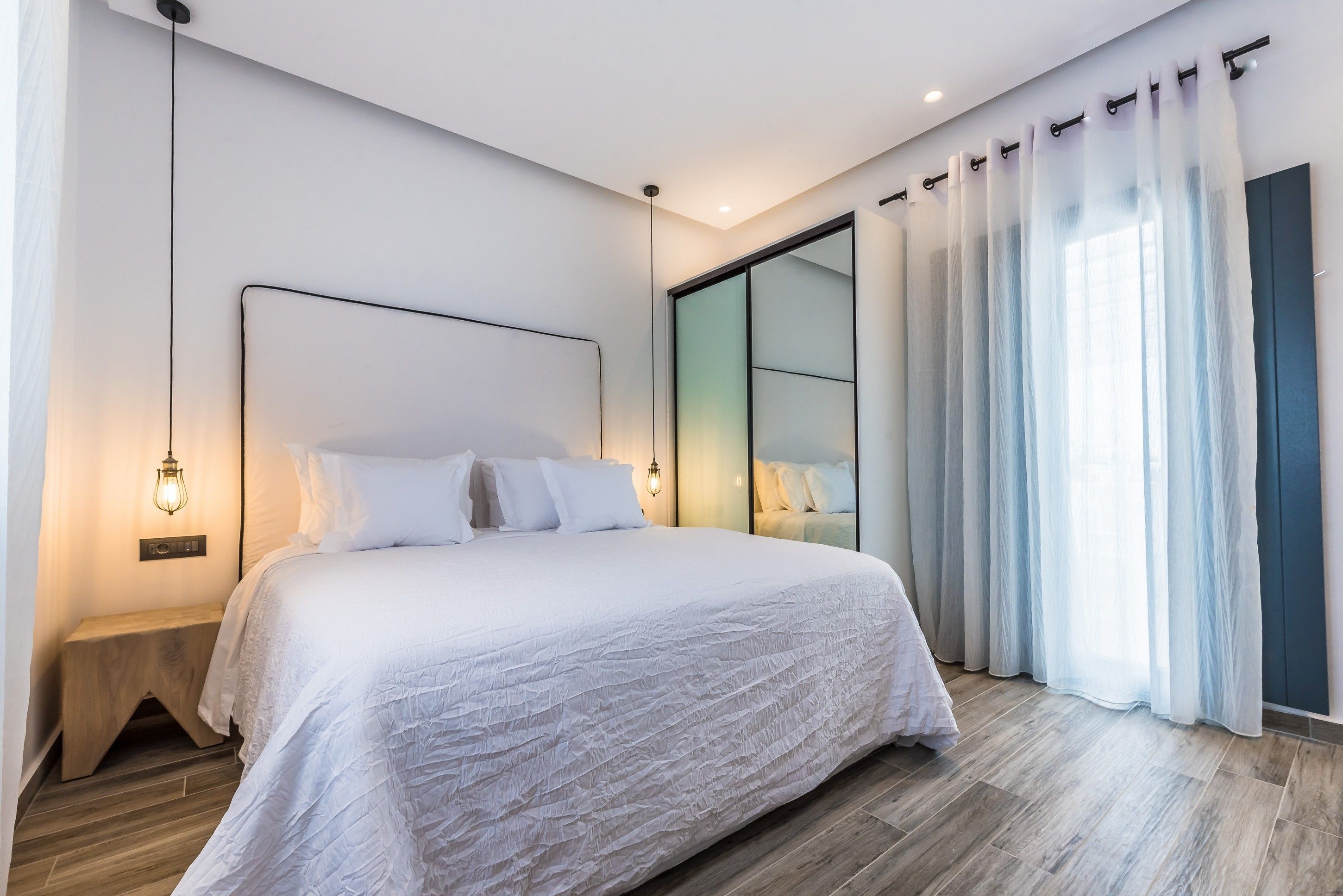 Un dormitorio moderno con una cama grande, una mesita de noche, una lámpara de pared y una puerta corrediza de vidrio con cortinas translúcidas.
