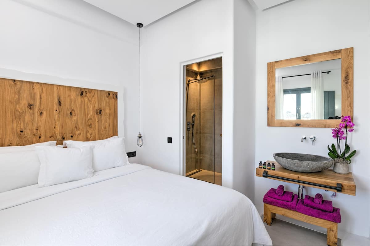 Een moderne slaapkamer met een houten hoofdbord, een groot bed, een douche met glazen wand, een houten wastafelmeubel met een waskom en een spiegel.