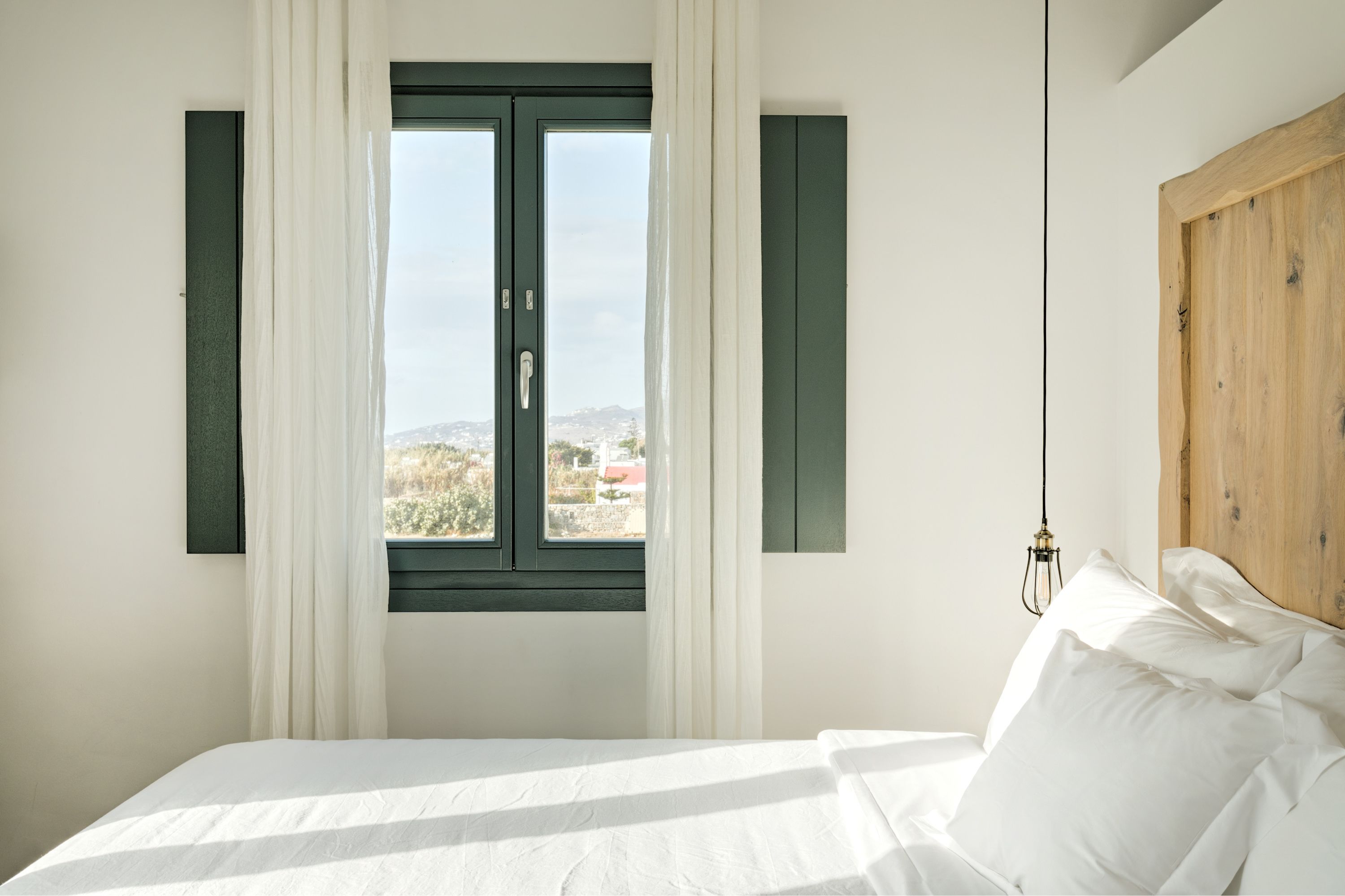 A bedroom with a bed, a window with curtains, a view of the outside, and a wooden headboard.