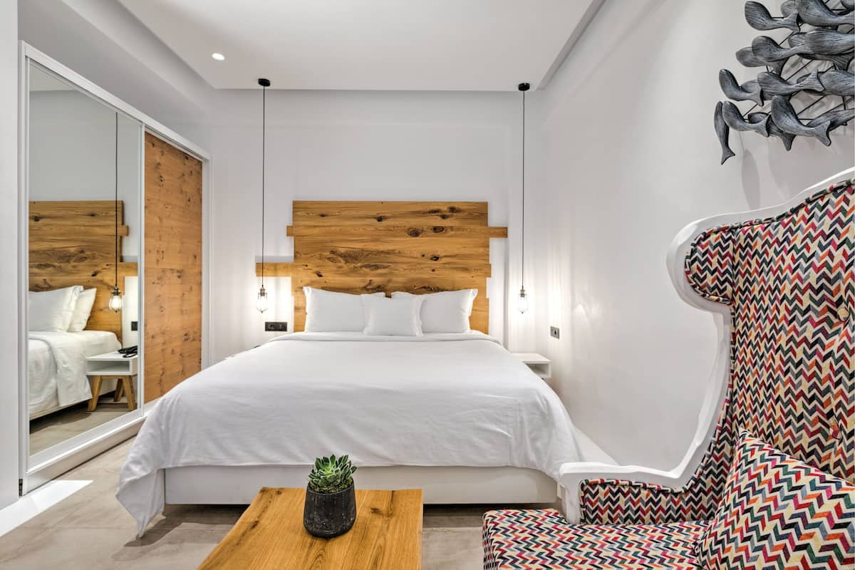 Een moderne slaapkamer met een houten hoofdbord, een wit bed, een kleurrijke fauteuil en een kleine tafel met een plant.