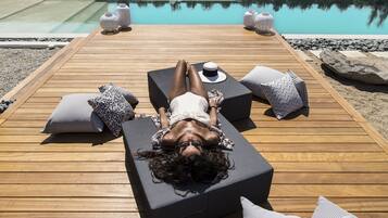 A person lying on a lounge chair by a poolside with a wooden deck, surrounded by pillows and a hat.