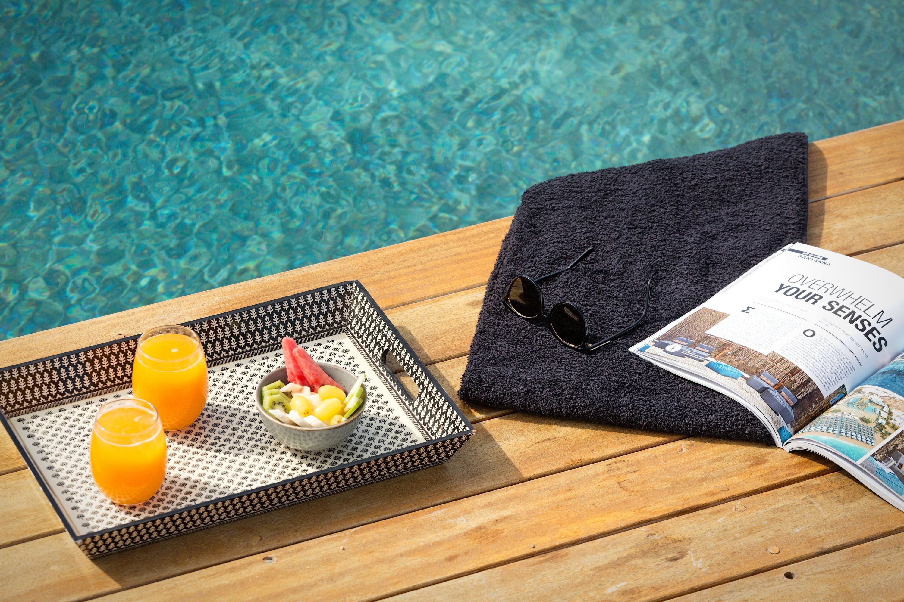 Ein Tablett mit zwei Gläsern Orangensaft, einer Obstschale, einem gefalteten Handtuch und einer Zeitschrift auf einem Holztisch an einem Pool.