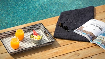 A tray with two glasses of orange juice and a bowl of fruit, a folded towel, and a magazine on a wooden table by a pool.
