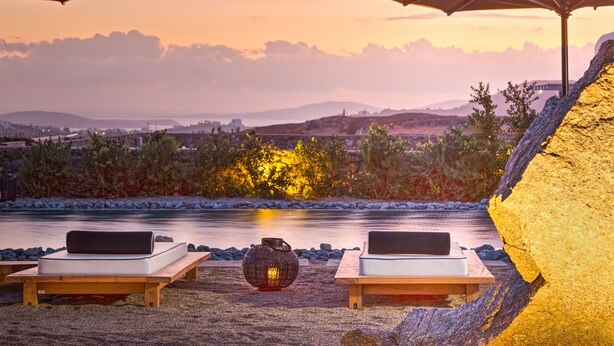 A serene outdoor seating area with two lounge chairs and a table, overlooking a body of water and a distant cityscape during sunset. - Portes Mykonos Suites & Villas (Mykonos)